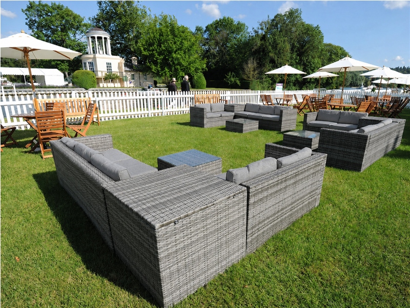Grey outdoor rattan sofas and tables arranged on a lawn, with wooden chairs, white parasols and a white picket fence in the background.