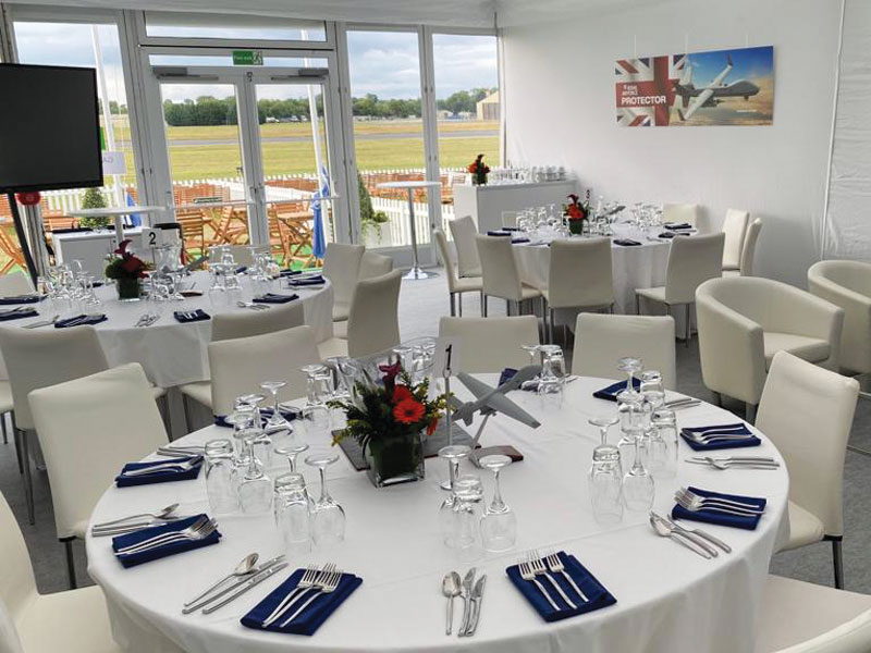 Round dining tables set with white tablecloths, glassware, blue napkins and red floral centrepieces in a bright event room with white chairs and large windows.