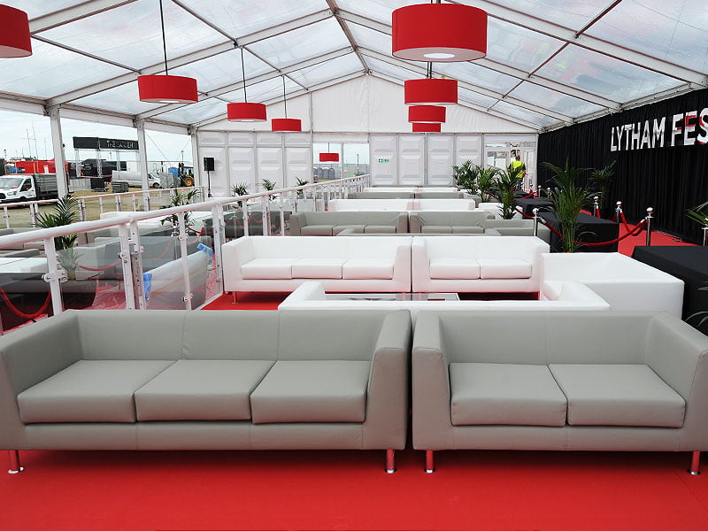 Event lounge inside a large marquee with rows of white sofas on red flooring, red pendant lights overhead, potted plants and a roped-off black stage area.