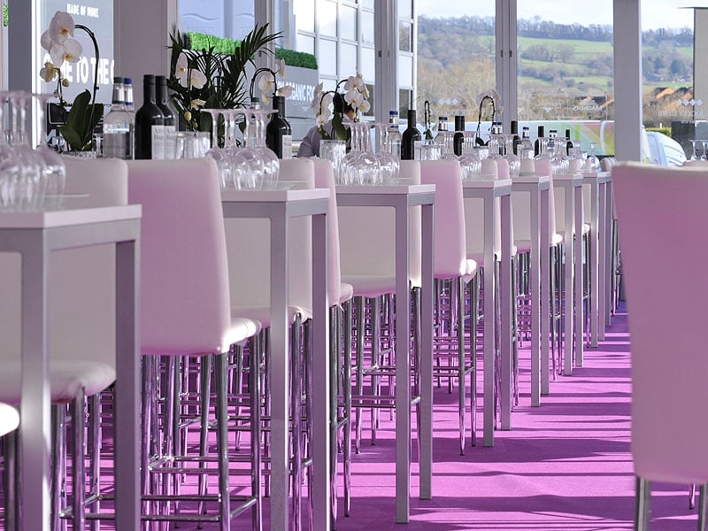 Bright modern bar or dining area with rows of white high tables and stools, glassware, bottles and white orchids, lit with pink-purple floor lighting beside large windows.