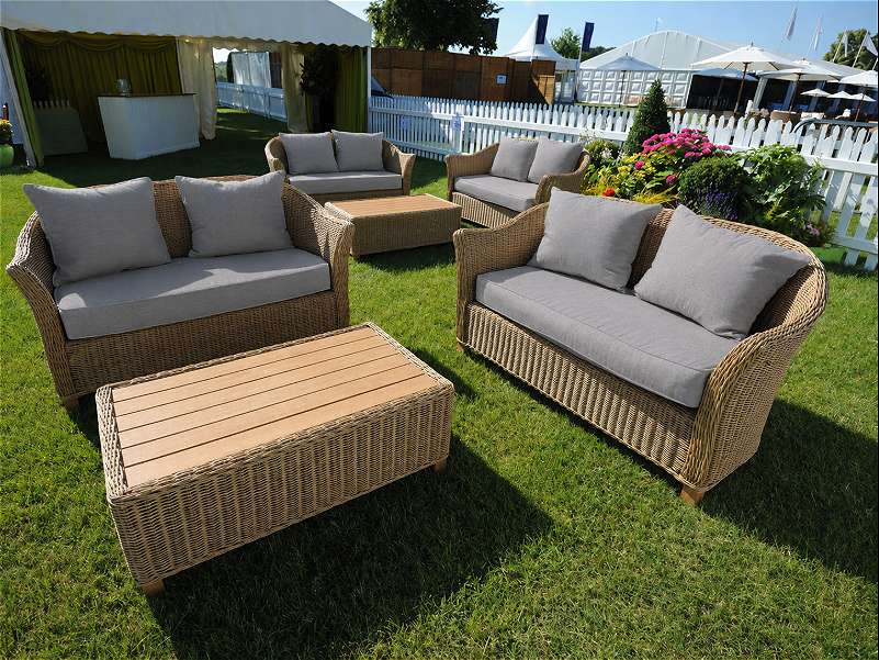 Wicker outdoor sofas with grey cushions and matching low tables arranged on grass beside colourful flower beds, white fencing and event marquees.