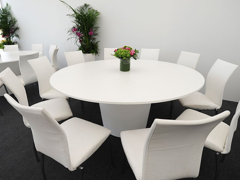 Large round white table surrounded by white upholstered chairs, with a small floral centrepiece in a bright indoor event space.