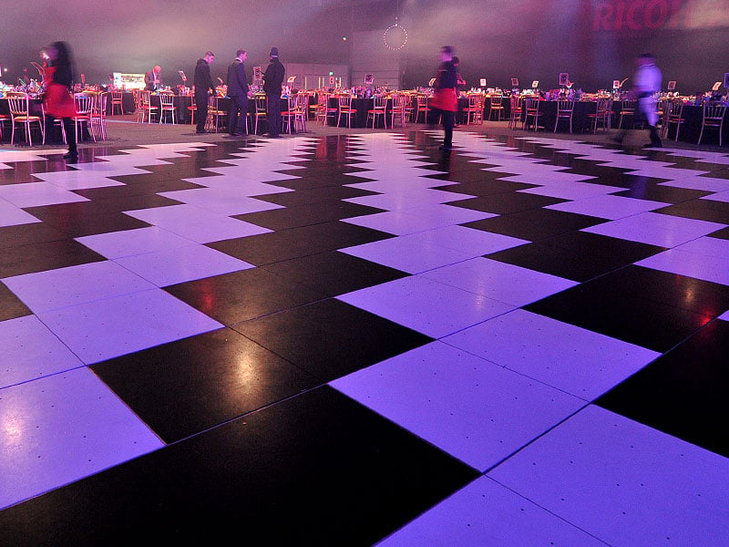 Large black-and-white chequered dance floor under purple lighting, surrounded by banquet tables and blurred figures in a spacious event venue.