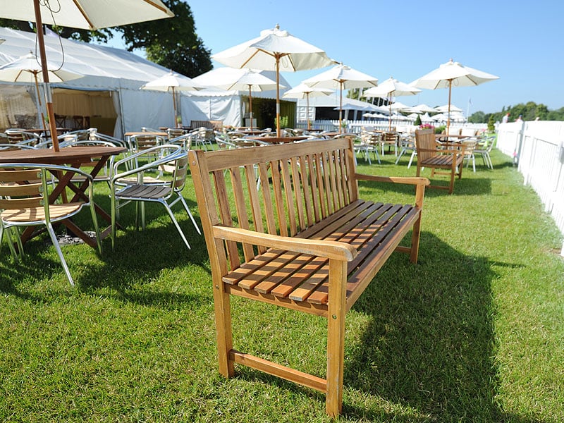 Wooden bench on a grassy outdoor hospitality area surrounded by café tables, metal chairs, white parasols and marquees beneath a clear blue sky.