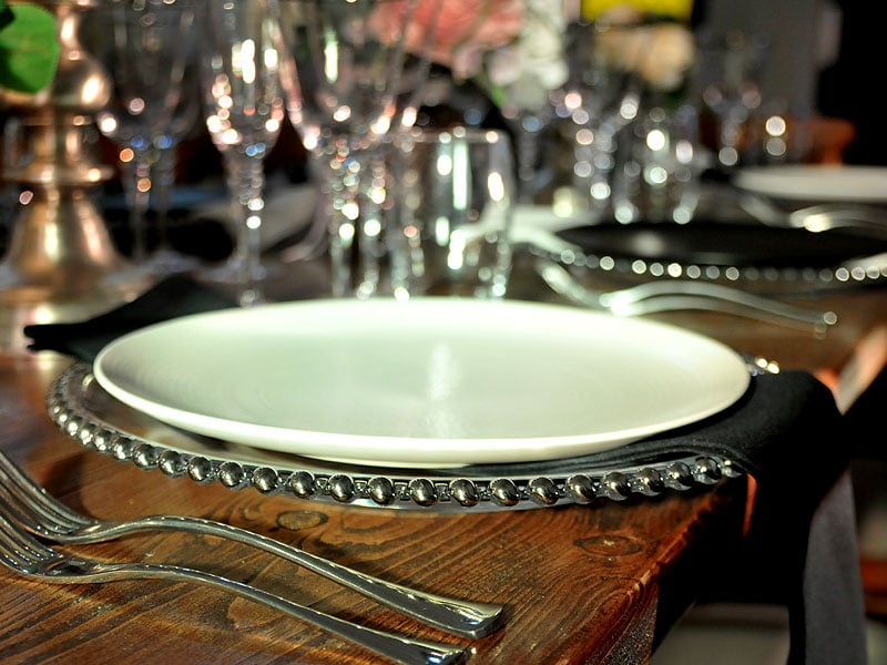 White dinner plate on a silver beaded charger with polished cutlery, black napkins and glassware on a wooden table.