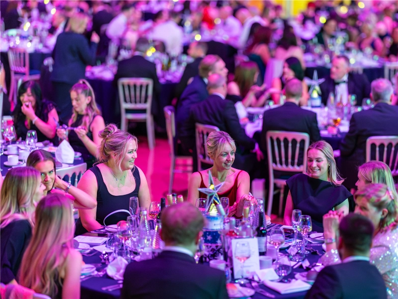 Guests in formal attire seated around crowded banquet tables, talking and smiling beneath vivid pink and purple event lighting.