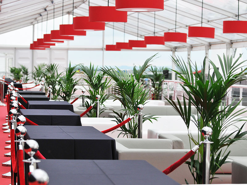 Marquee lounge with rows of black-covered tables and white sofas separated by red ropes, surrounded by potted palms beneath red pendant lights.