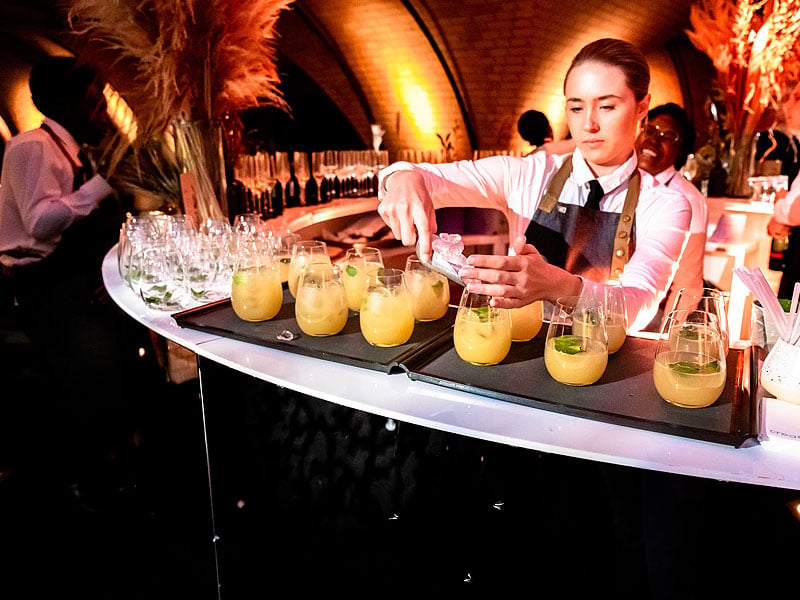 Bartender preparing yellow cocktails in stemless glasses at a curved bar, surrounded by glassware under warm orange lighting.