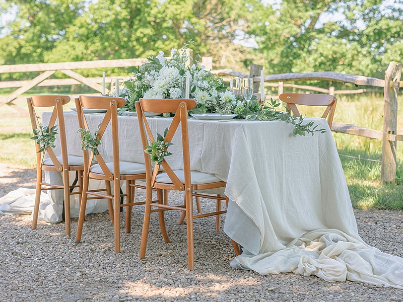 Outdoor dining table draped in white linen, surrounded by wooden chairs and decorated with white flowers, greenery and candles beside a rustic fence.
