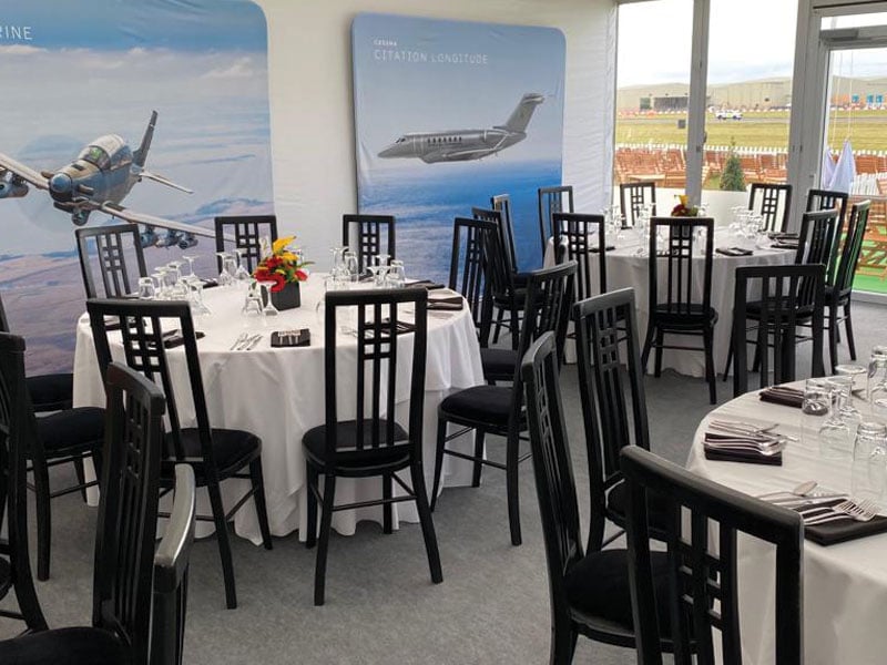 Round dining tables with white tablecloths, black chairs, glassware and place settings inside a bright event space, with large aircraft graphics on the walls and a view of an airfield through the windows.