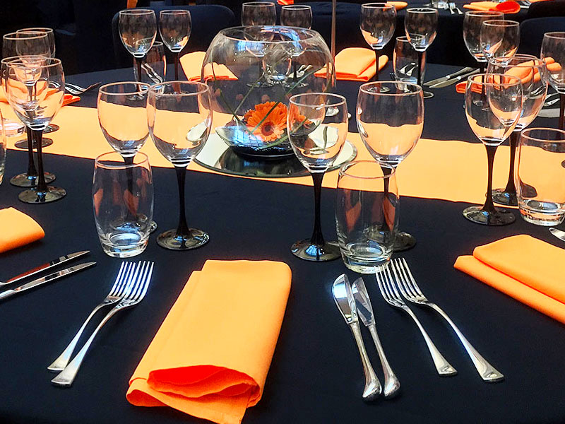 Formal table setting with black linen, bright orange napkins, polished cutlery, numerous wine glasses and a glass bowl containing orange flowers.