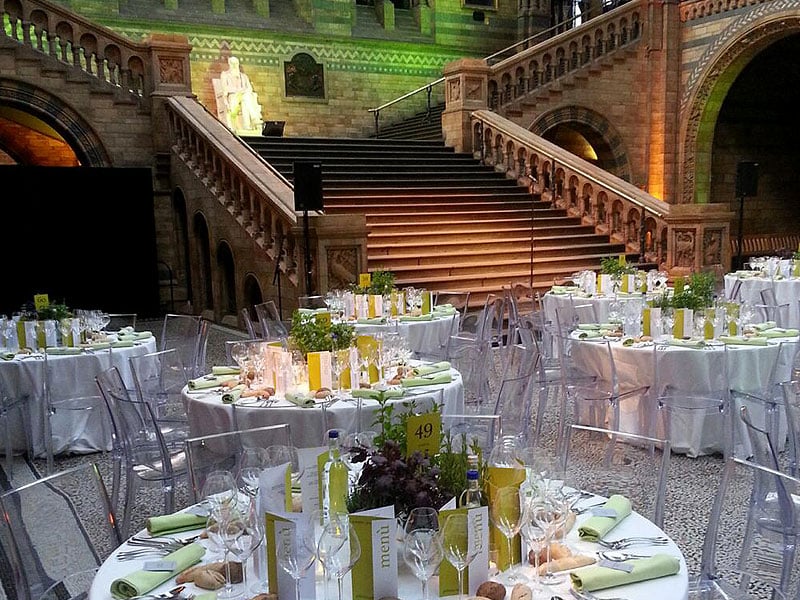 Formal dining tables with white tablecloths, green napkins and clear chairs arranged in a grand stone hall beneath a wide staircase.
