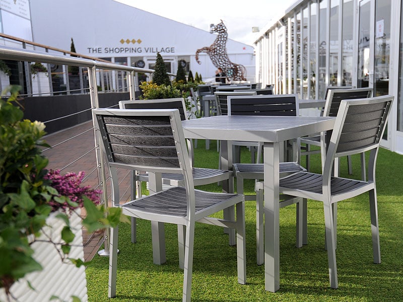 Grey metal tables and chairs on artificial grass outside a shopping village, with planters and a large horse sculpture in the background.