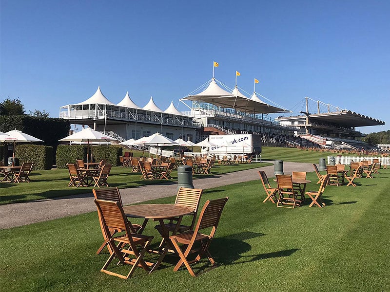 Wooden tables and chairs arranged on a large lawn beside a grandstand and white peaked canopies under a clear blue sky.
