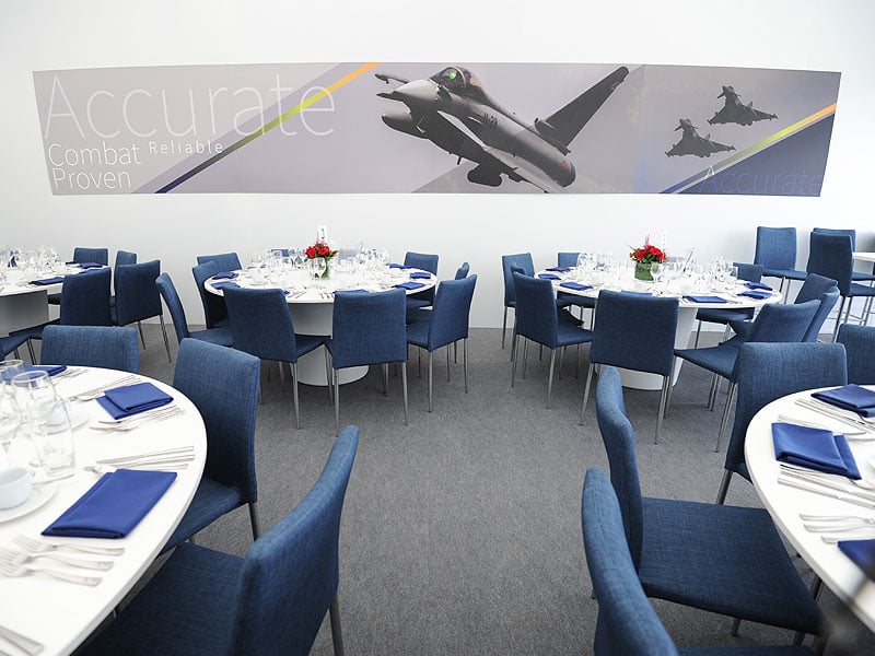 Formal dining room with round white tables set with glassware, blue napkins and red floral centrepieces, surrounded by blue chairs beneath a large fighter jet mural.