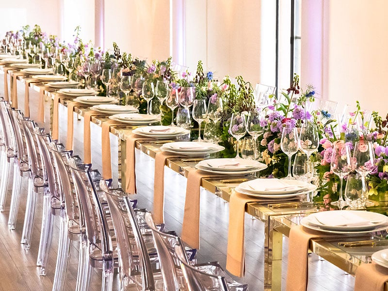 Long mirrored banquet table set with white plates, numerous wine glasses and beige napkins, bordered by clear chairs and a continuous arrangement of pink, purple and green flowers.