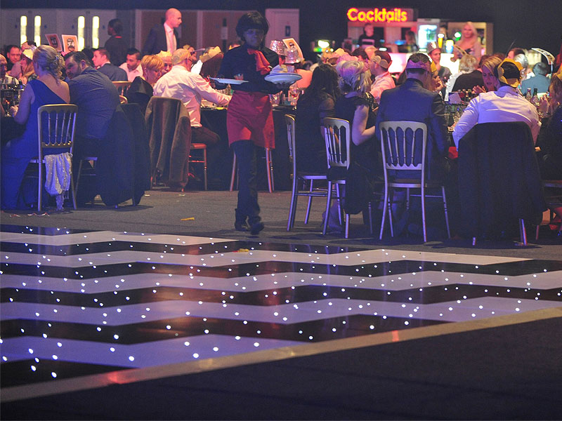 Guests dine at round tables during a dimly lit event, while a server carries plates beside an illuminated black-and-white dance floor.