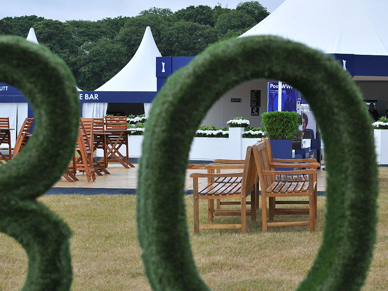 Large green topiary loops frame wooden outdoor chairs and tables on a lawn, with white marquee tents and trees in the background.