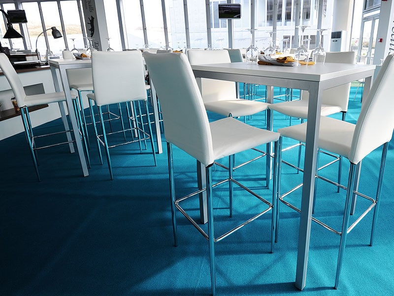 Bright modern dining area with tall white chairs around square tables on vivid turquoise carpet, surrounded by floor-to-ceiling windows.