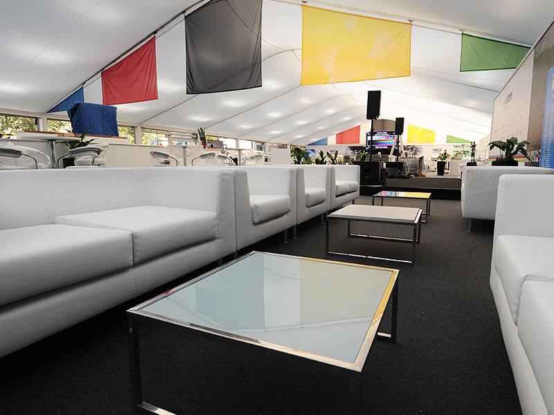 White sofas and glass-topped coffee tables arranged inside a large marquee, with colourful fabric panels hanging from the ceiling and a small stage at the far end.