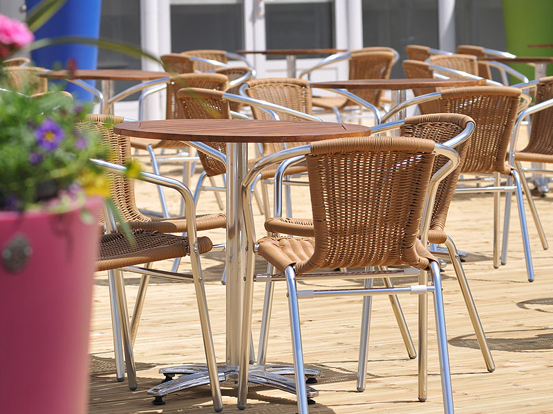Outdoor café terrace with wicker chairs and wooden-topped tables on decking, with a blurred flower planter in the foreground.