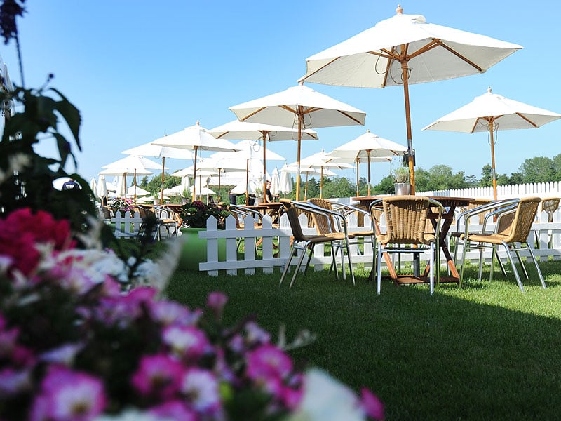 Outdoor dining area on a lawn with wicker chairs, wooden tables and white parasols, bordered by a low white picket fence and blurred pink flowers in the foreground.