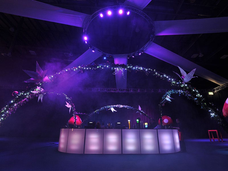 Illuminated bar beneath large floral arches and a circular lighting rig, with purple stage lighting and decorative bird figures overhead.