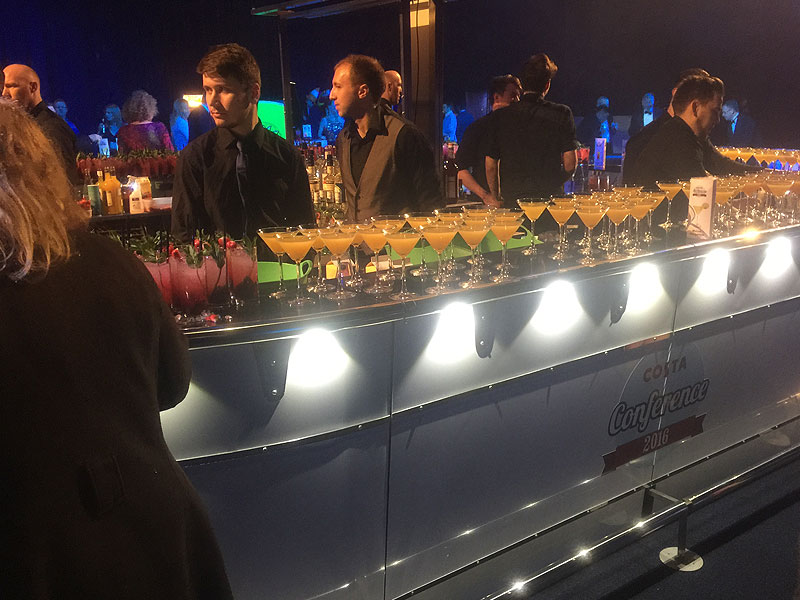 Bartenders stand behind an illuminated event bar lined with rows of cocktails, with guests gathered in the dimly lit background.