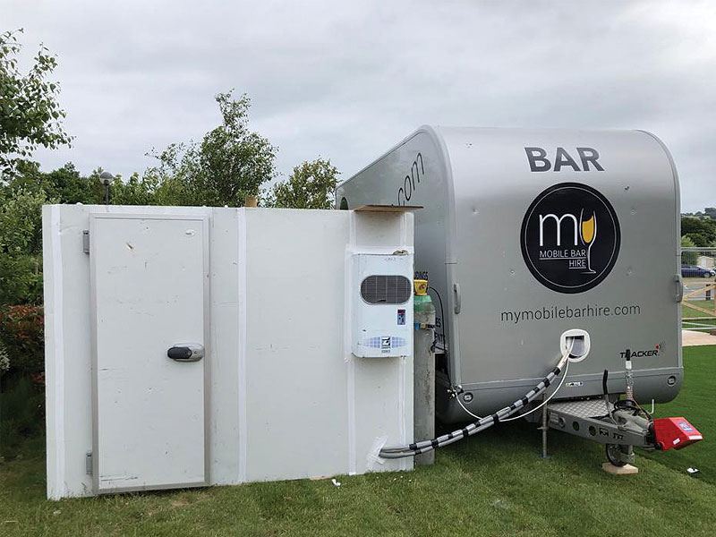 Silver mobile bar trailer on grass beside a white screened enclosure, with “BAR” and “mymobilebarhire.com” visible on the trailer.