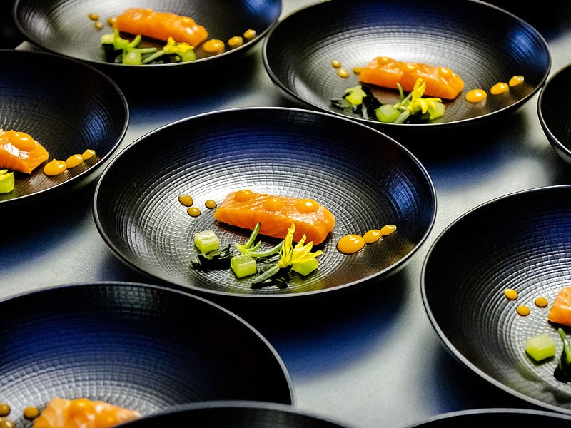 Multiple black bowls arranged for plating, each containing a piece of orange fish with green vegetables and dots of orange sauce.