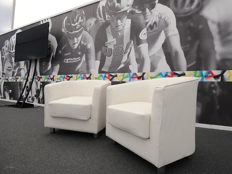 Two white armchairs beside a black display screen, set against a large monochrome mural of racing cyclists.