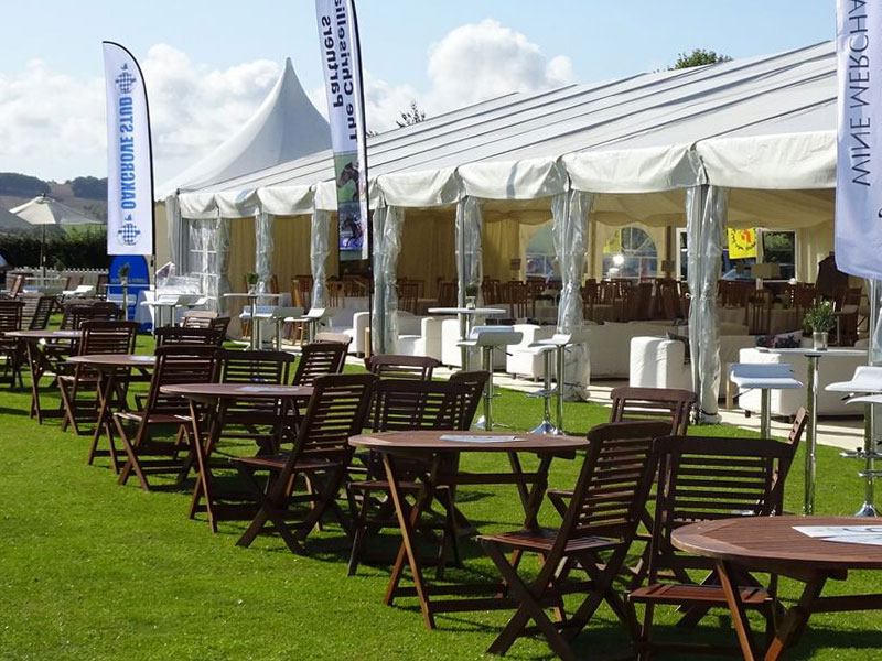 Outdoor event seating with wooden tables and chairs on grass beside a large white marquee, with white lounge furniture and tall flags nearby.