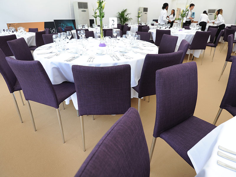 Round dining tables with white tablecloths, purple upholstered chairs, glassware and tall floral centrepieces in a bright event space.