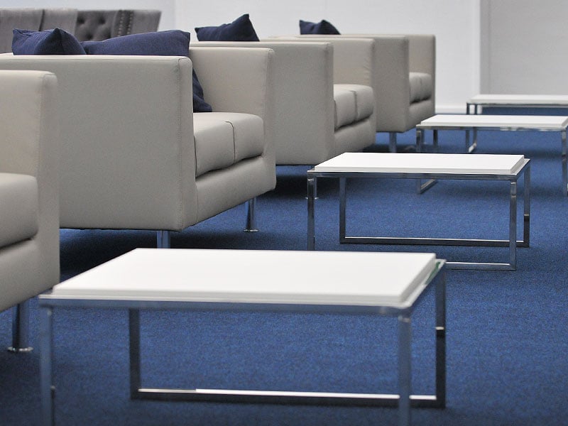 Grey lounge chairs with dark cushions arranged beside low white tables on a blue carpet.