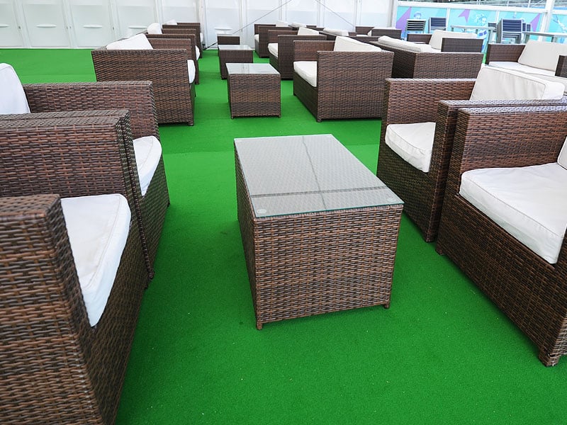 Lounge seating area with brown wicker armchairs and sofas, white cushions and glass-topped tables arranged on bright green flooring.