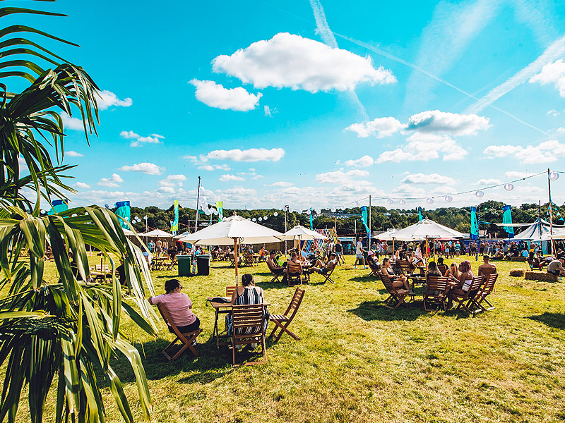 Outdoor festival seating area on grass with wooden tables and chairs, large white parasols, string lights and groups of people beneath a bright blue sky.