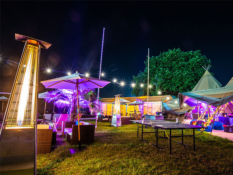 Colourfully lit outdoor event space at night with patio heaters, parasols, lounge seating, tents and a table-tennis table on grass.
