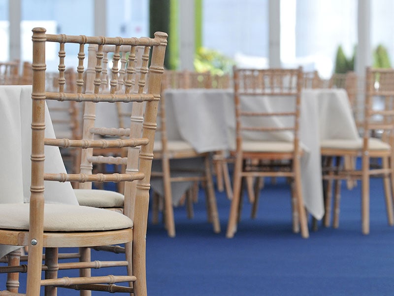 Light wooden banquet chairs with cream seat cushions arranged around white-clothed tables on a blue carpet in a bright event space.