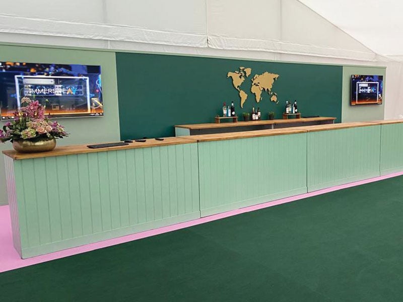 Long mint-green bar with a wooden counter inside a marquee, backed by a dark green wall featuring a gold world map, bottles and two wall-mounted screens.