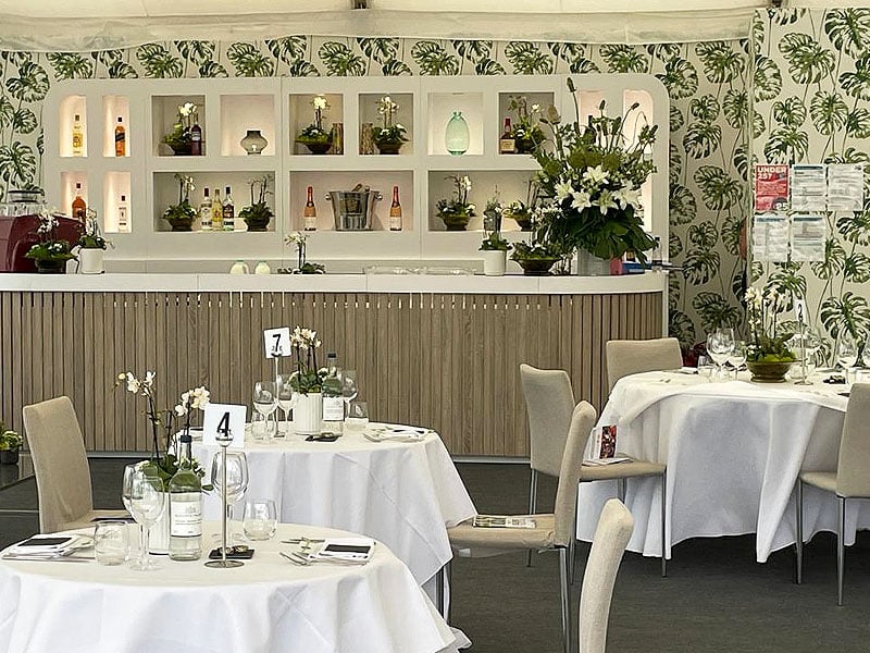 Bright event dining area with round, white-clothed tables, numbered place settings, floral centrepieces and a bar backed by tropical leaf-patterned walls.