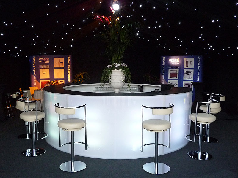 Illuminated circular bar surrounded by white stools beneath a black, star-lit ceiling, with a large floral arrangement at the centre.