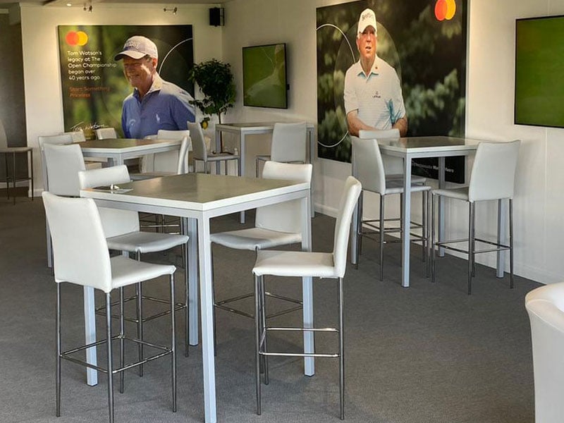 Bright hospitality lounge with white high tables and stools, wall-mounted screens, and large golf-themed posters featuring a male golfer and Mastercard branding.