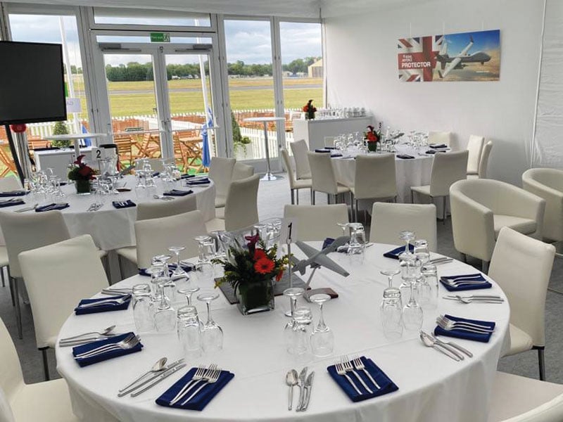 Bright event dining room with round, white-clothed tables set with glassware, navy napkins and red floral centrepieces, surrounded by white chairs beside large windows overlooking an outdoor seating area and airfield.
