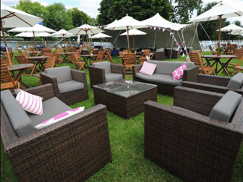 Outdoor event seating area on grass, with dark wicker sofas and armchairs around a glass-topped table, surrounded by wooden tables, chairs and white parasols.