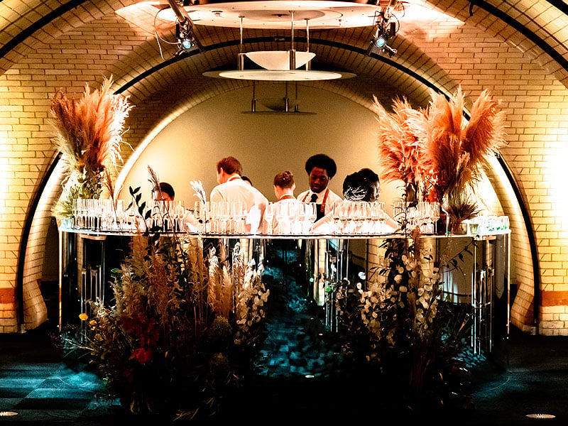 Staff prepare drinks behind a mirrored bar beneath a brick arch, framed by tall dried grasses, floral arrangements and rows of glasses.