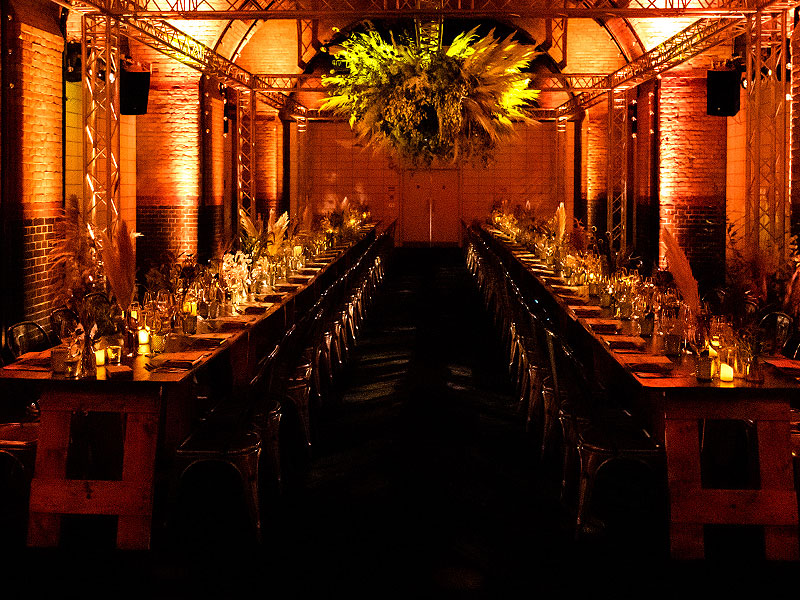 Two long banquet tables set with glassware and candles beneath a large suspended floral arrangement, illuminated by warm orange lighting in a brick hall.