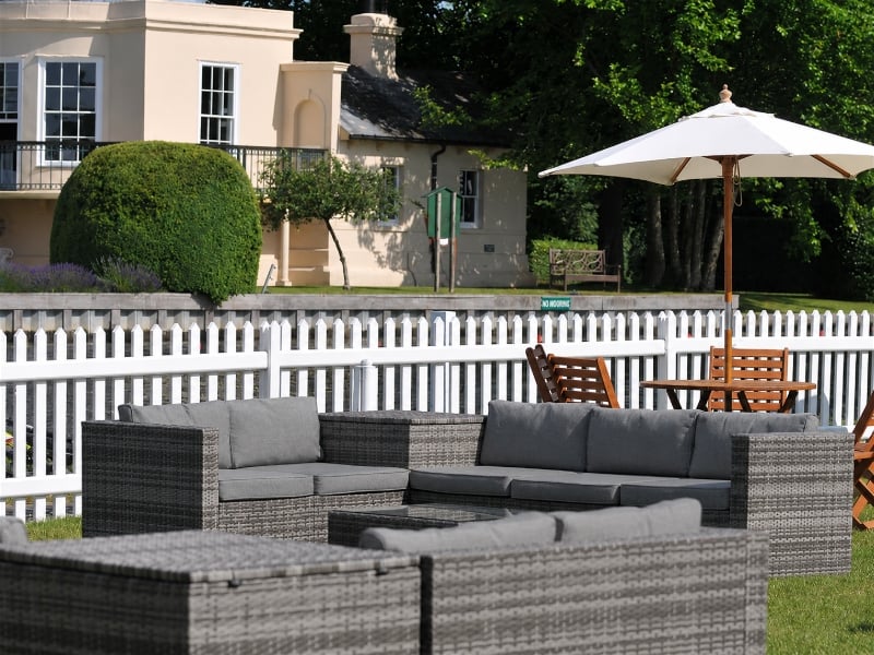 Grey rattan outdoor sofas and a wooden table beneath a white parasol on a lawn, enclosed by a white picket fence with a cream building and trees behind.