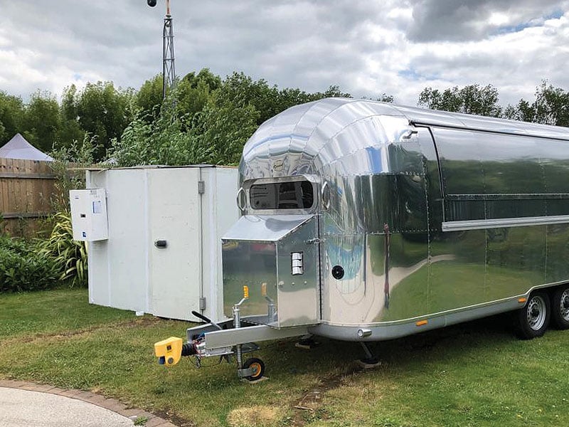 Polished silver catering trailer parked on grass beside a large white refrigeration unit, with trees and cloudy skies behind.