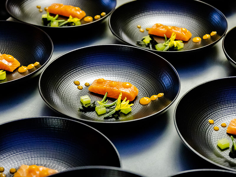 Black bowls containing neatly plated orange fish, green vegetables and small dots of orange sauce.