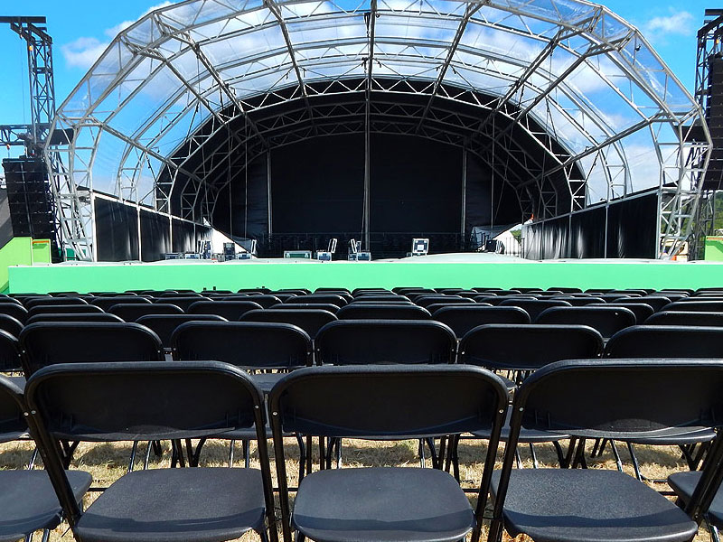 Rows of black folding chairs face a large covered outdoor stage with metal trussing and a bright green front panel beneath a blue sky.